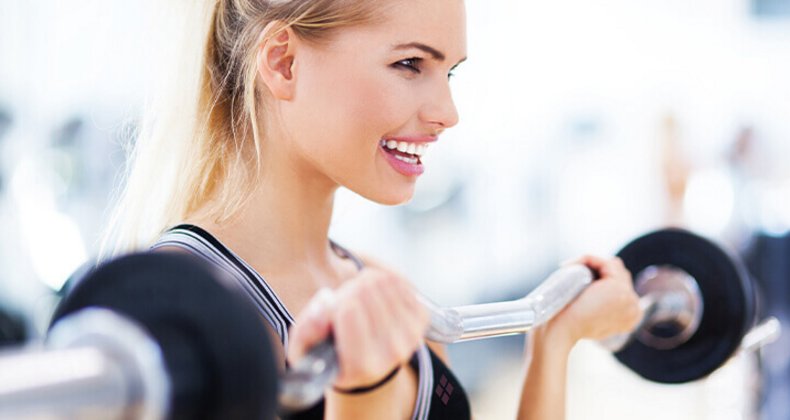  Eine lächelnde, sportliche Frau hebt eine Langhantel in einem hellen Fitnessstudio.